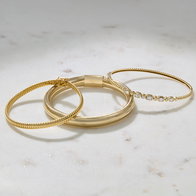 Three gold bracelets, including two textured gold bangles and one gold bangle with bezel‑set diamonds, arranged on a marble surface.