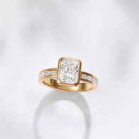 Bezel-set diamond engagement ring with a yellow gold band and baguette diamond accents displayed on a light background.